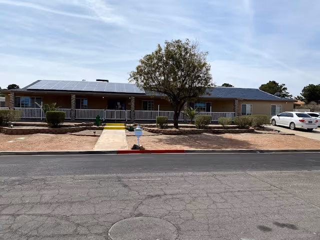 Single-story building with a front porch, solar panels on the roof, a tree and shrubs in the front yard, and a white car parked on the right side driveway under a partly cloudy sky.