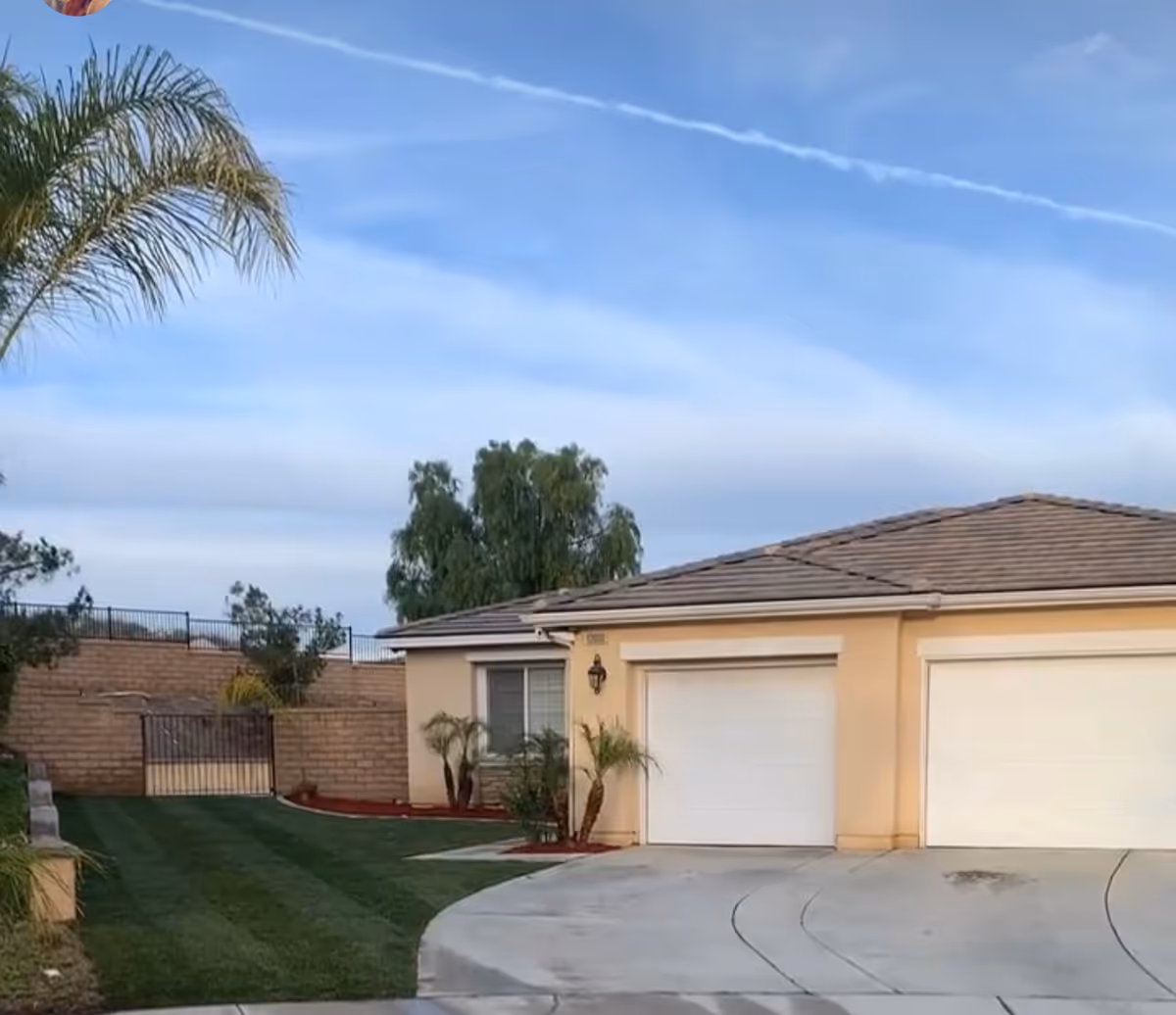 Exterior view of a single-story residential building with a tiled roof, two white garage doors, a small window, and a driveway. The front yard has neatly trimmed grass, small palm trees, and a brick wall with a metal gate. The sky is clear with some wispy clouds.