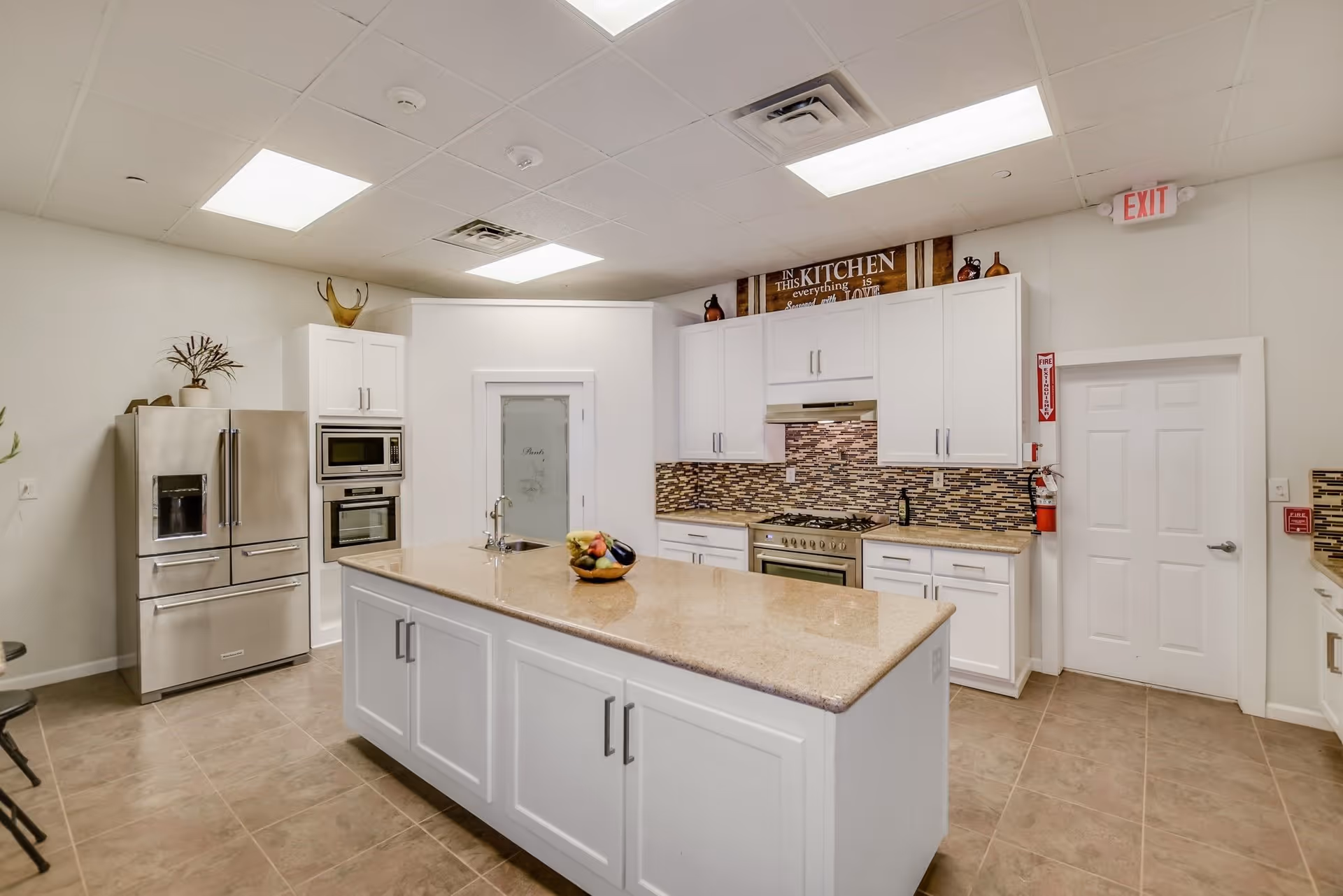Bright assisted-living community kitchen with a central island, stainless steel refrigerator, oven, white cabinets, and a tiled backsplash.