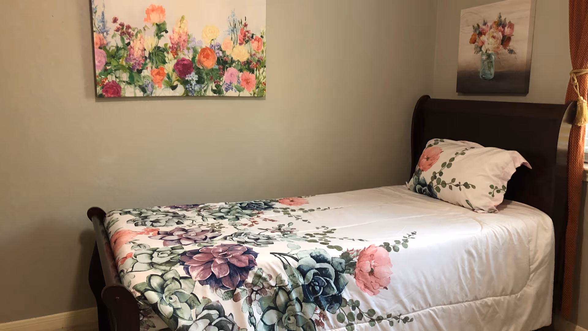 A neatly made single bed with floral-patterned bedding and pillow in a small bedroom. The bed has a dark wooden frame. Two floral paintings hang on the beige walls above and beside the bed.