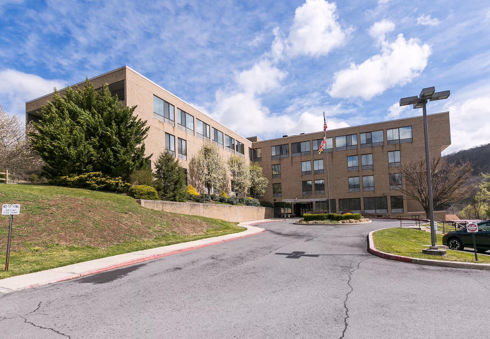 Exterior view of a multi-story brick building with many windows, surrounded by greenery and a driveway leading to the entrance. There are two flagpoles with flags in front of the building under a partly cloudy sky.