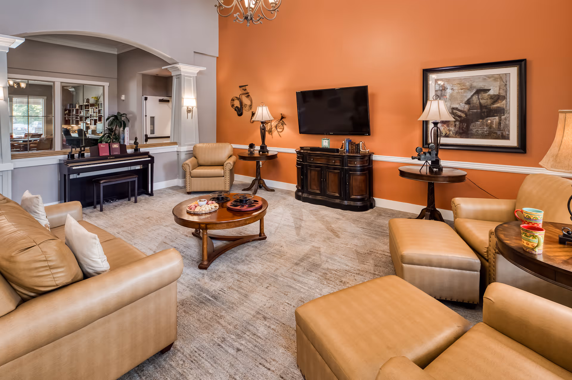 Comfortable communal living room with tan leather seating, a round coffee table, wall-mounted TV, piano and an orange accent wall.