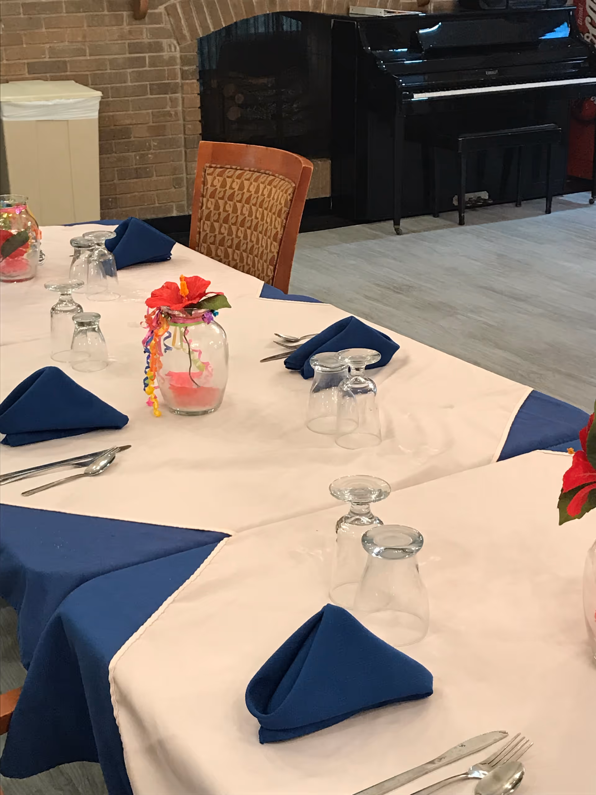 Table set for dining with blue napkins, inverted glassware and floral jar centerpieces in a room with a piano in the background.