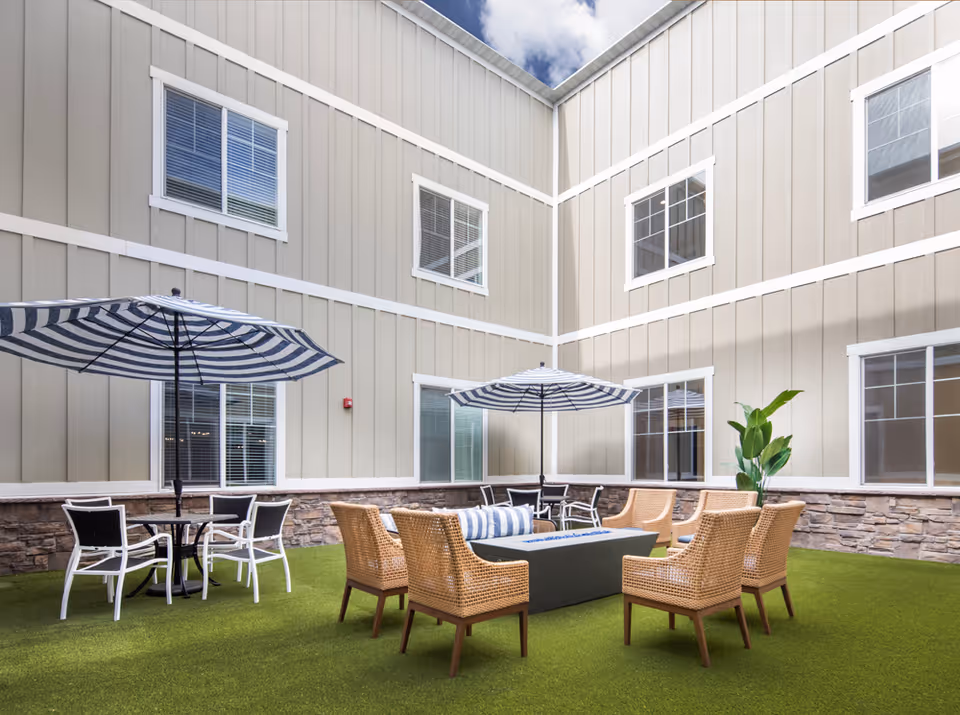 Outdoor courtyard area with artificial grass, surrounded by beige building walls with white trim and multiple windows. The space features two seating areas with striped umbrellas, one with a round table and white chairs, and the other with wicker chairs around a rectangular fire pit table. A potted plant is visible near the wall.