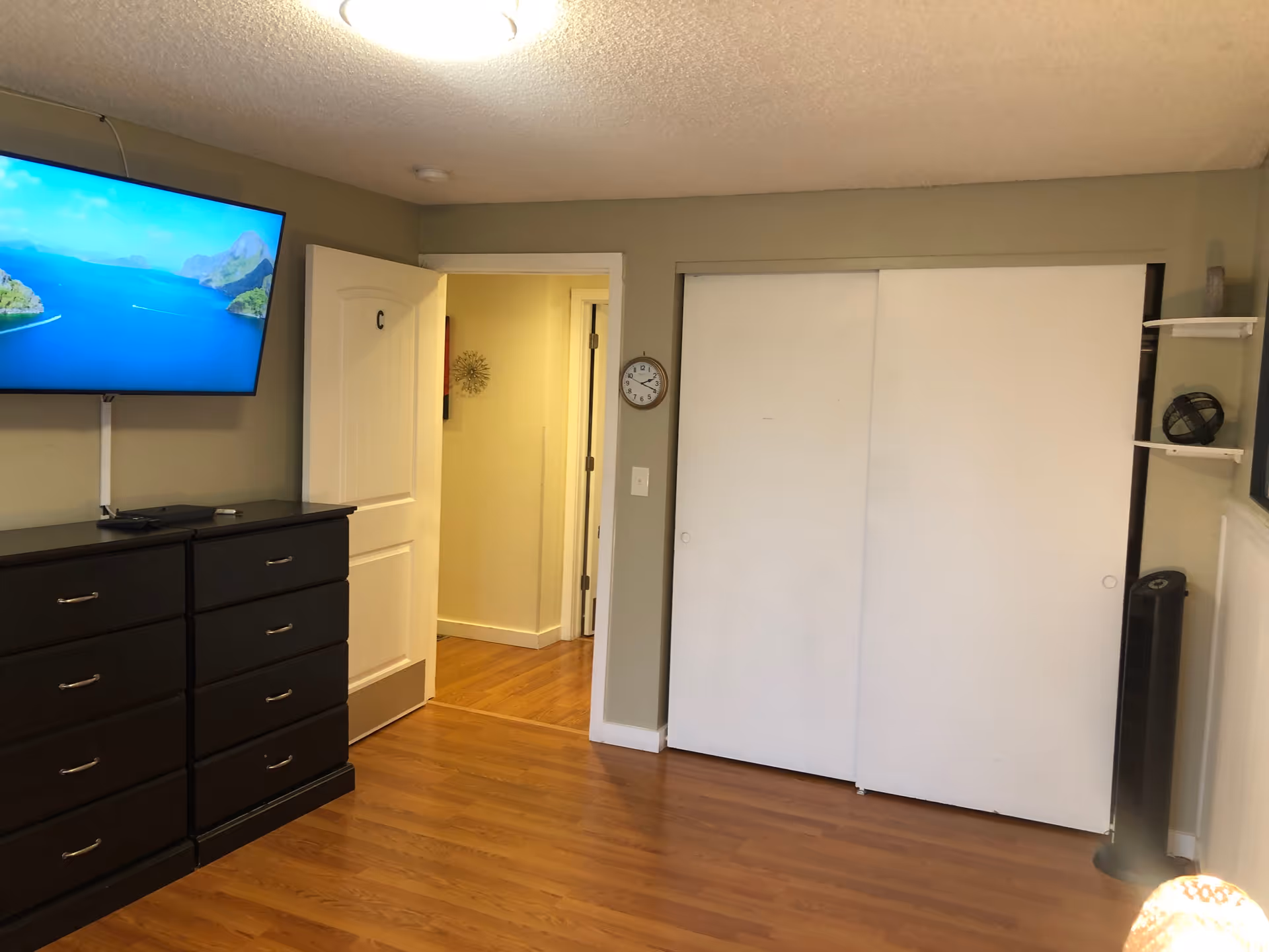 Interior view of a room with wooden flooring, a wall-mounted flat screen TV displaying a scenic ocean view, a dark wooden dresser with multiple drawers, a white door labeled with the letter 'C' slightly open revealing a hallway, a wall clock, and a large white sliding closet door. There is also a black tower fan and small shelves with decorative items on the right side.