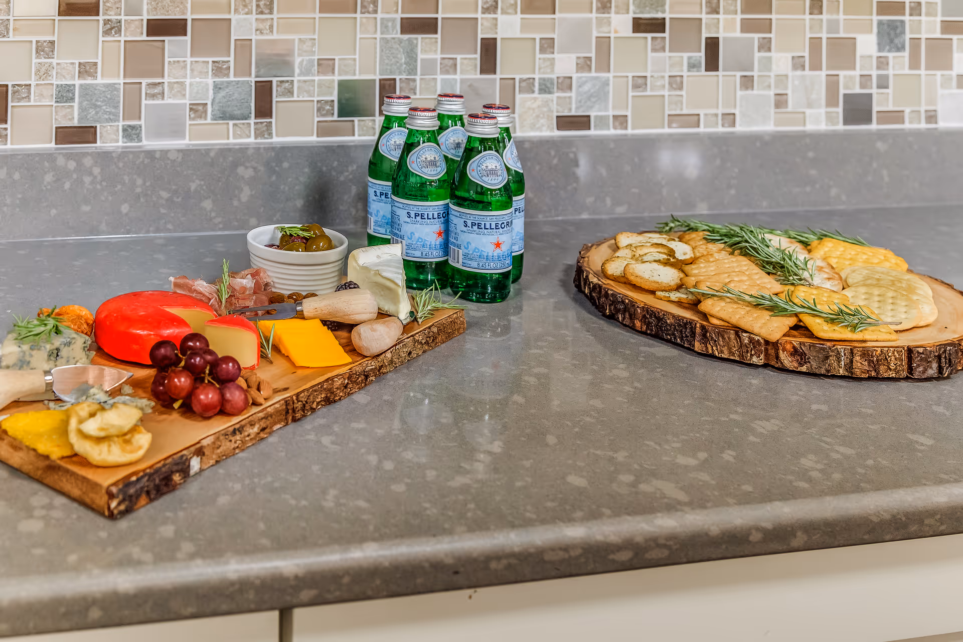 Two wooden serving boards with cheeses, crackers, grapes and bottles of S.Pellegrino on a kitchen countertop with a mosaic tile backsplash.