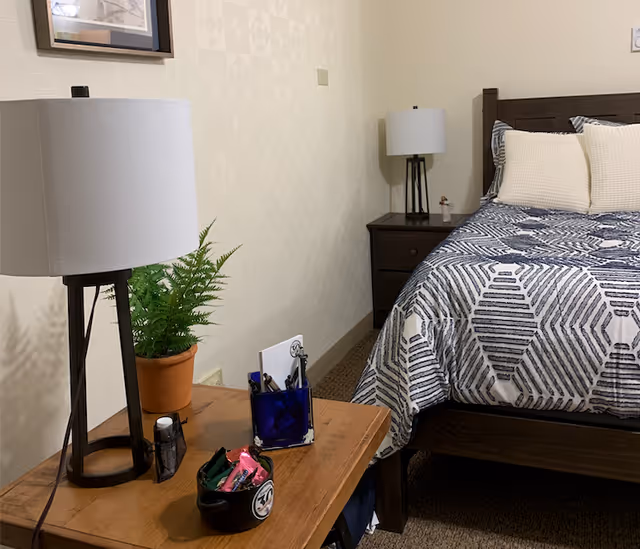 A bedroom with a wooden bed frame and a bed covered with a blue and white patterned comforter and cream-colored pillows. There is a wooden side table with a lamp, a potted fern, a blue pen holder, and a small black container with items on it. Another wooden nightstand with a lamp and a small figurine is visible next to the bed.