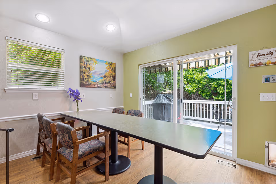 Dining area with a long rectangular table and upholstered wooden chairs facing sliding glass doors that open to a patio.