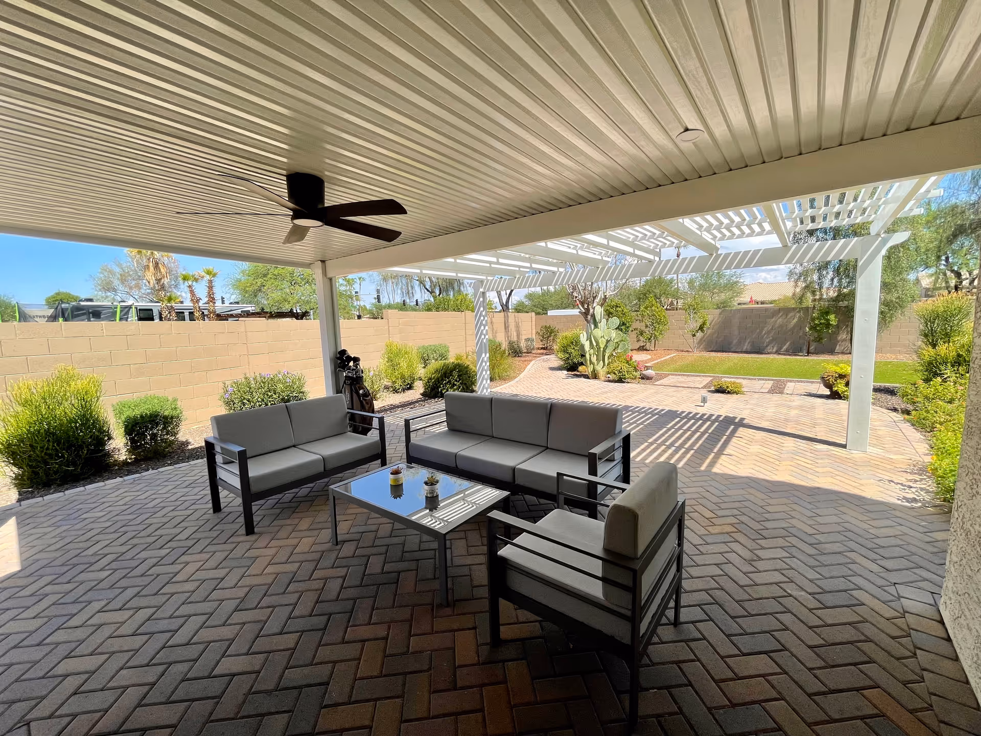 Outdoor patio area with a covered seating space featuring a ceiling fan, two sofas, and a chair around a glass-top coffee table. The patio has brick flooring and overlooks a garden with bushes, trees, and a cactus under a pergola.