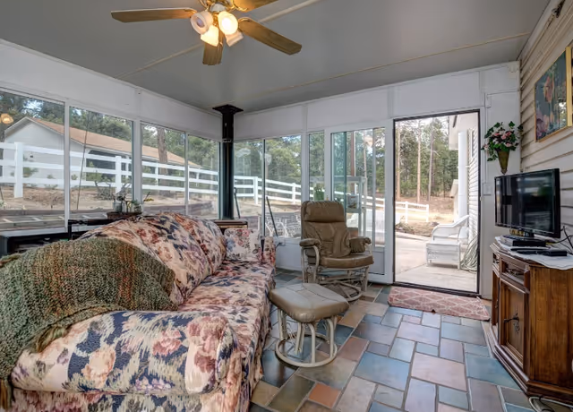 A cozy sunroom with large windows and sliding glass doors showing an outdoor view with trees and a white fence. The room features a floral-patterned sofa with a green knitted throw, a beige recliner chair with matching footrest, a ceiling fan with lights, a wood stove, and a wooden cabinet with a flat-screen TV on top. The floor is tiled with multicolored square tiles.