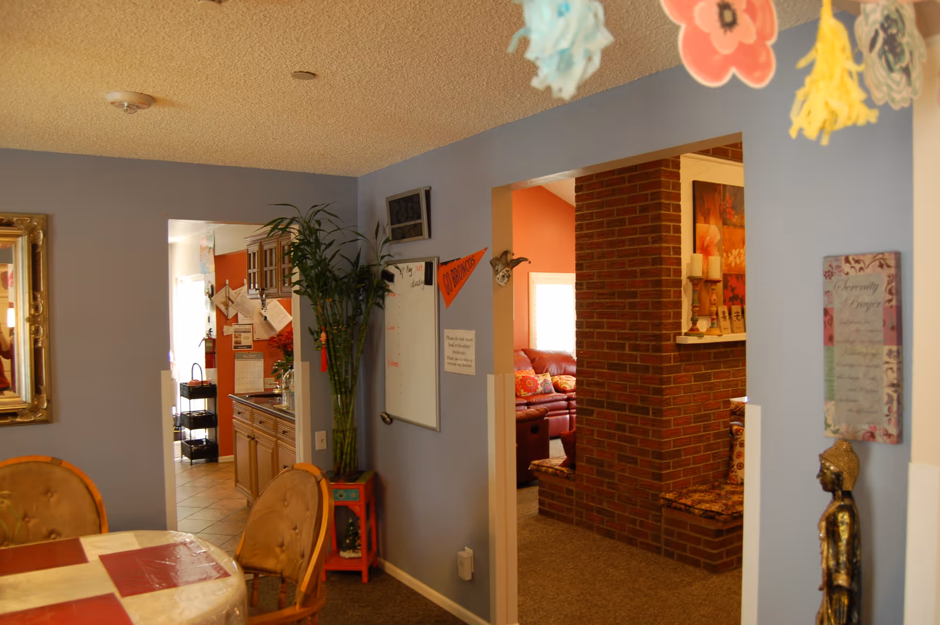 Interior view of an assisted living facility showing a dining area with a round table and chairs, a blue wall with a whiteboard and decorations, a tall plant in a red stand, and an adjacent room with a brick fireplace and a red couch with colorful pillows.