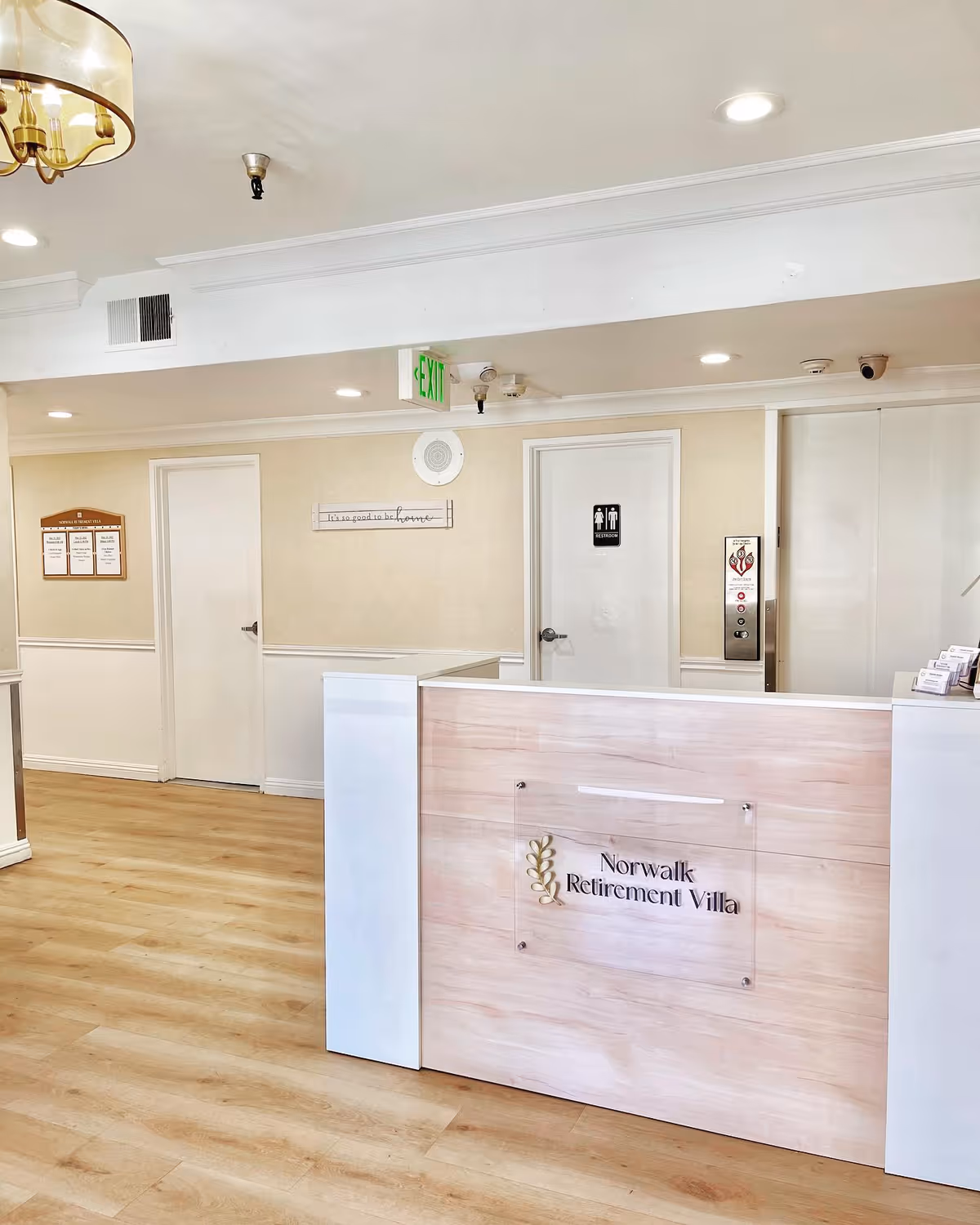 Reception area of Norwalk Retirement Villa with a light wood and white front desk, beige walls, wooden floor, and ceiling lights. There is a door with a restroom sign and an elevator next to it. A green exit sign is visible above the door.