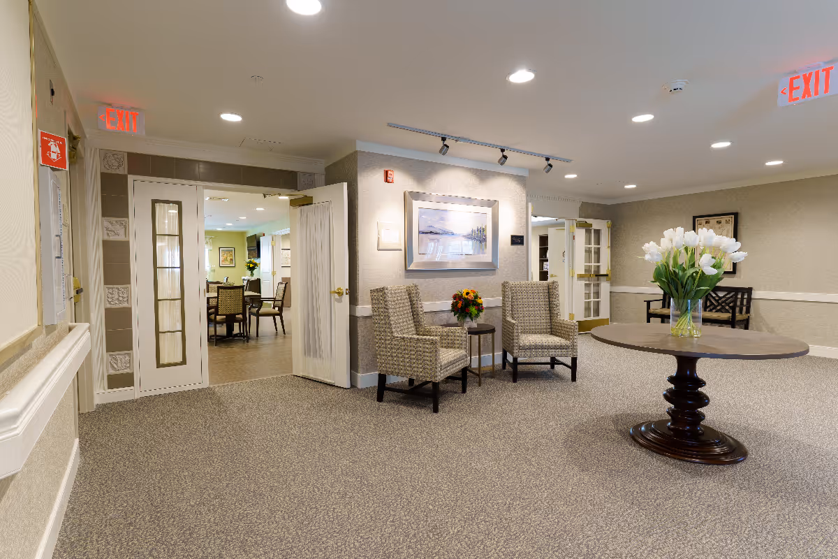 A well-lit interior common area in a senior living facility with a round wooden table holding a vase of white flowers in the center. Two patterned armchairs with a small table and a flower arrangement are placed against the wall beneath a framed landscape painting. Double doors lead to a dining area with tables and chairs. The walls are beige with white trim, and there are exit signs visible on the ceiling.