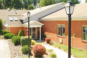 Exterior view of a single-story brick building with a sloped roof, featuring a small covered entrance with white columns. The surrounding area includes landscaped bushes, a stone pathway, and two black lamp posts. Trees are visible in the background.