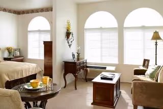A bright and cozy living space with a bed on the left, a small round table with breakfast items, a wooden dresser, a coffee table, a sofa, and large arched windows with blinds letting in natural light.