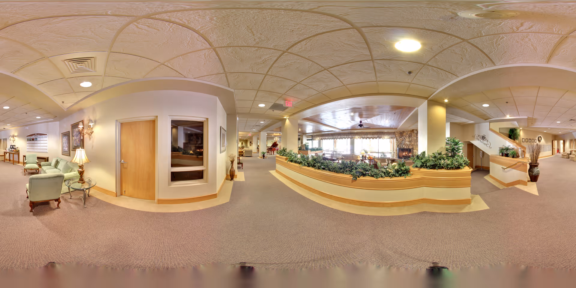 Wide panoramic view of a senior living facility interior featuring a spacious hallway with beige carpeting and cream-colored walls. On the left, there is a sitting area with light green upholstered chairs, a glass-top side table with a lamp, and framed artwork on the walls. In the center, a raised planter with green plants separates the hallway from a dining area with large windows and a stone fireplace. To the right, there is a staircase leading to an upper floor and additional hallway space with decorative plants and wall art. The ceiling has a textured design with recessed lighting.