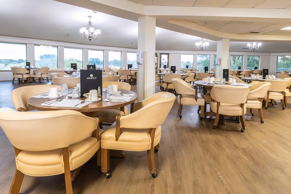 Spacious dining room with multiple round wooden tables surrounded by beige cushioned chairs on wheels. Each table is set with white cups, glasses, silverware, and a black menu labeled 'Asher Point'. Large windows line the walls, allowing natural light to fill the room, and chandeliers hang from the ceiling.