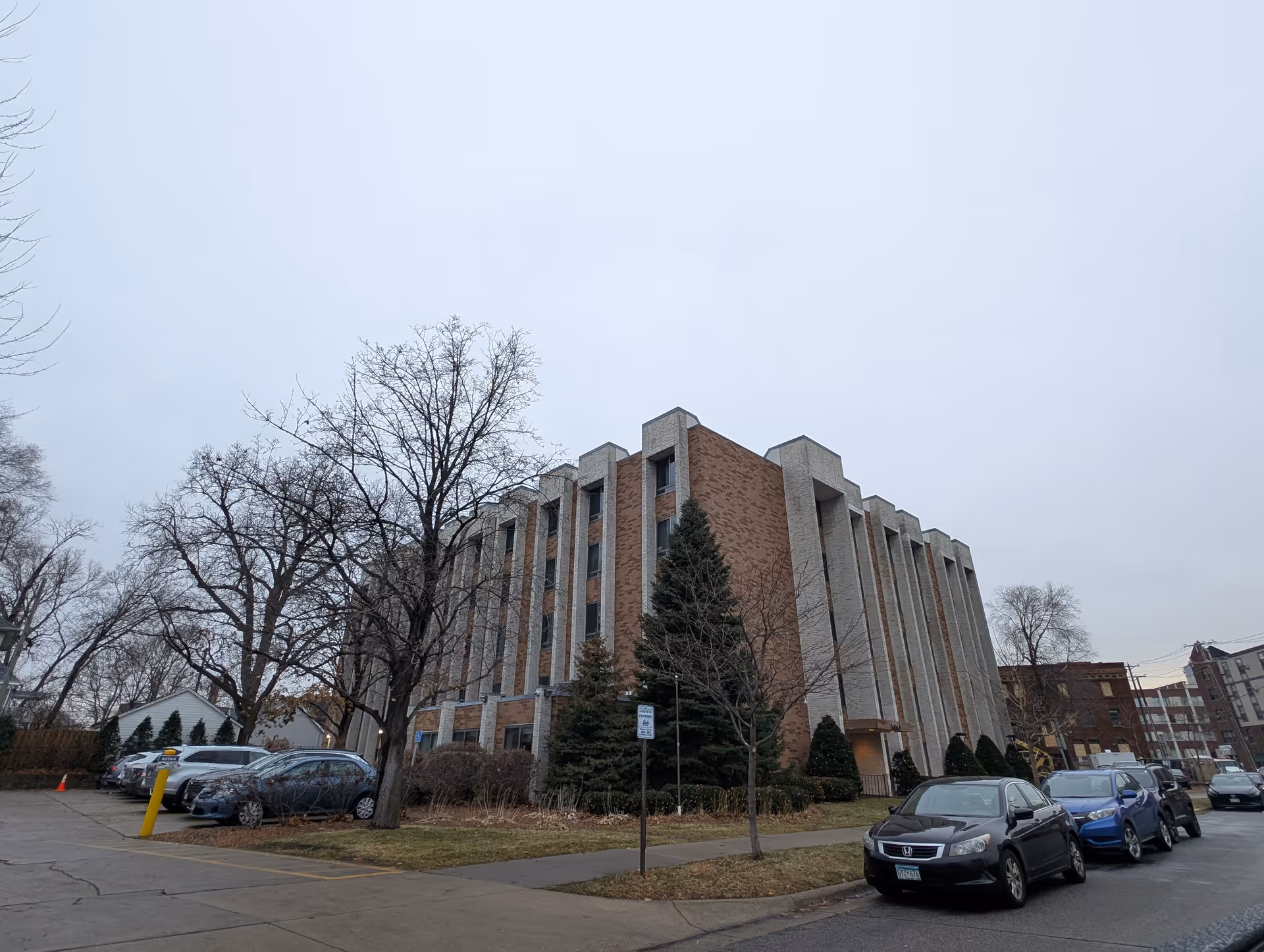 Exterior view of a multi-story brick and concrete building with several windows, surrounded by leafless trees and parked cars along the street and parking lot on a cloudy day.