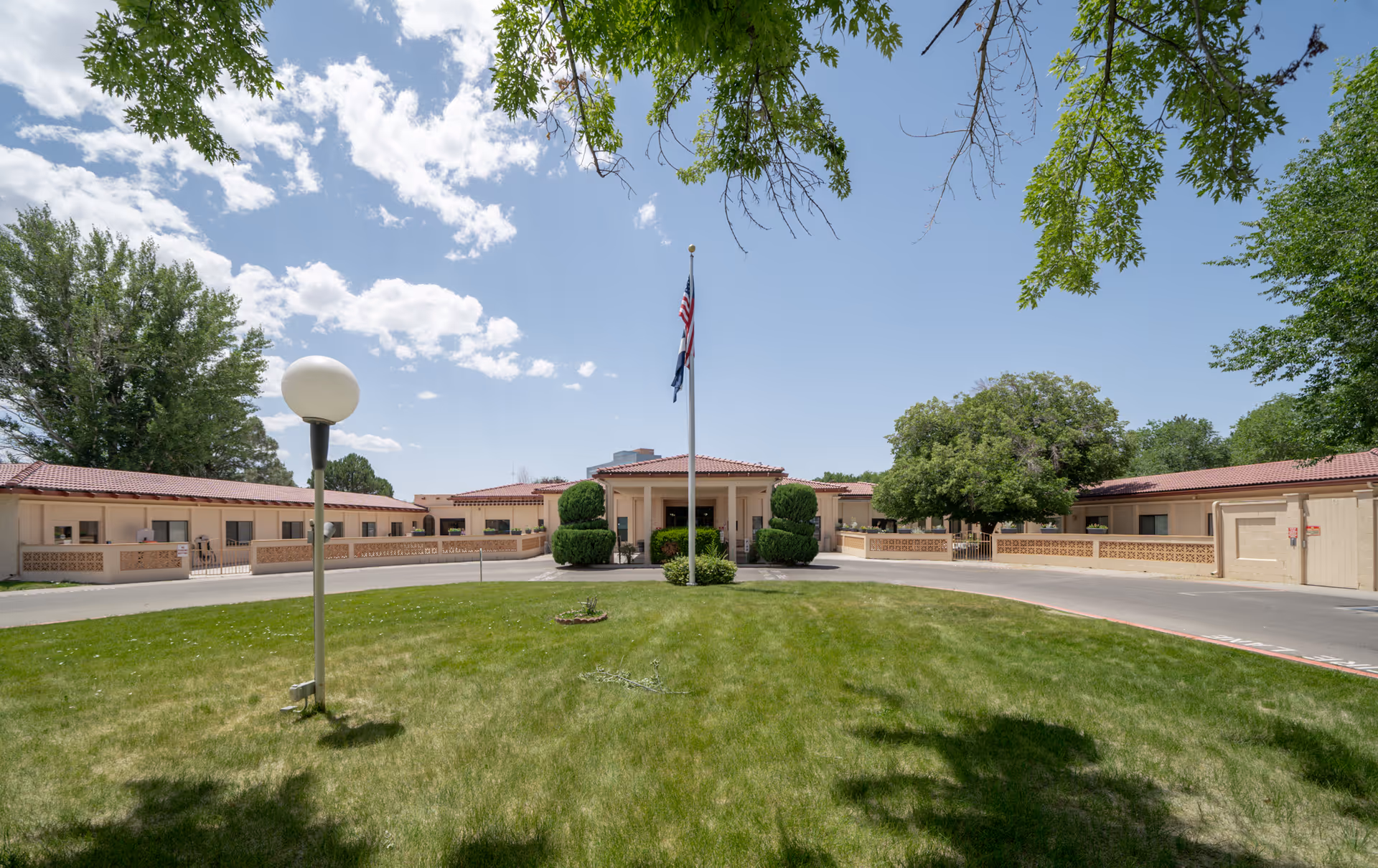 Front exterior view of La Villa Grande Care Center showing a single-story building with a red-tiled roof, a circular driveway, a flagpole with an American flag, green grass, and trees under a partly cloudy sky.