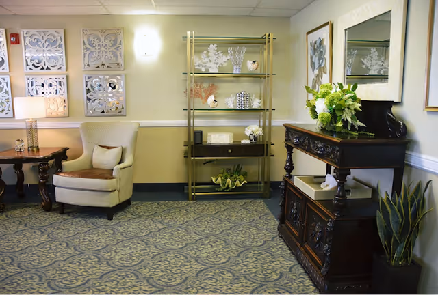 A cozy senior living facility common area with a beige armchair, a wooden side table with a lamp, decorative wall art, a gold and glass shelving unit with various ornaments, and a dark wooden console table with a floral arrangement and a large mirror above it. The room has patterned carpet and neutral-colored walls.