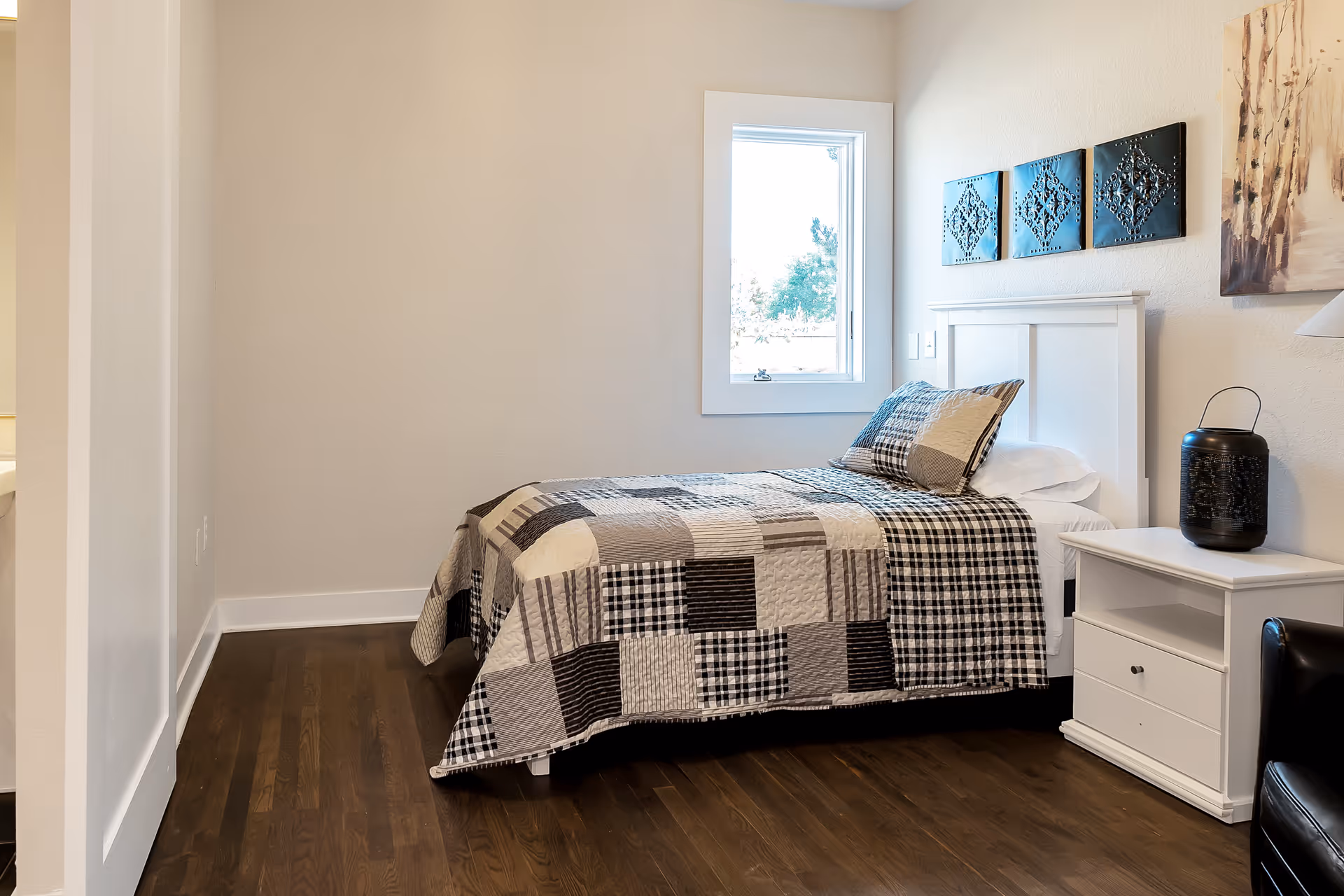A neatly made single bed with a patchwork quilt and matching pillow in a senior living bedroom. The room has light-colored walls, a window letting in natural light, a white nightstand with a decorative black lantern, and wall art above the bed.