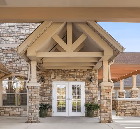 Entrance of a building with stone walls and a peaked roof supported by columns. There are double glass doors in the center with potted plants on either side. A pergola structure extends to the right side of the entrance.