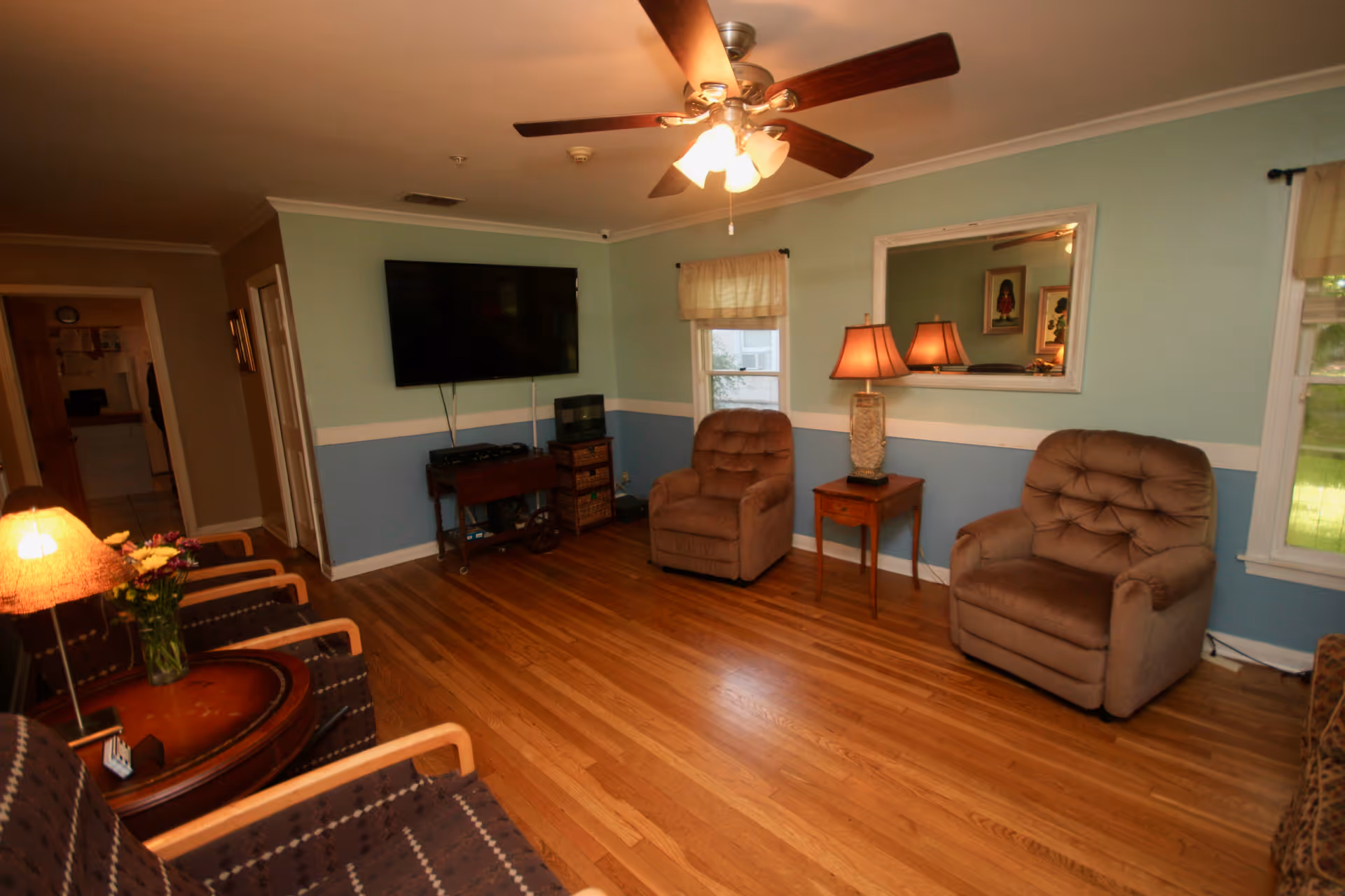 A cozy living room with wooden floors, two brown recliner chairs, two patterned armchairs, a round wooden side table with a lamp and flowers, a wall-mounted flat screen TV, a small wooden table with a lamp, and two windows with curtains. The walls are painted light blue with a white trim, and a ceiling fan with lights is on the ceiling.
