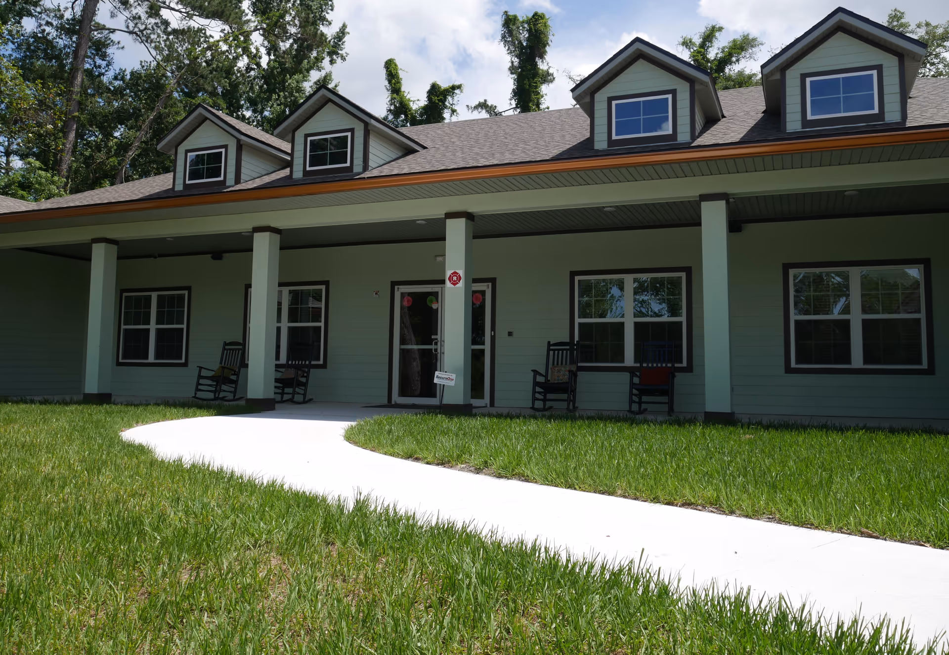 Front exterior view of a single-story building with a covered porch featuring four columns, four windows, and three dormer windows on the roof. There are four rocking chairs on the porch and a curved concrete walkway leading up to the entrance. The building is surrounded by green grass and trees in the background.