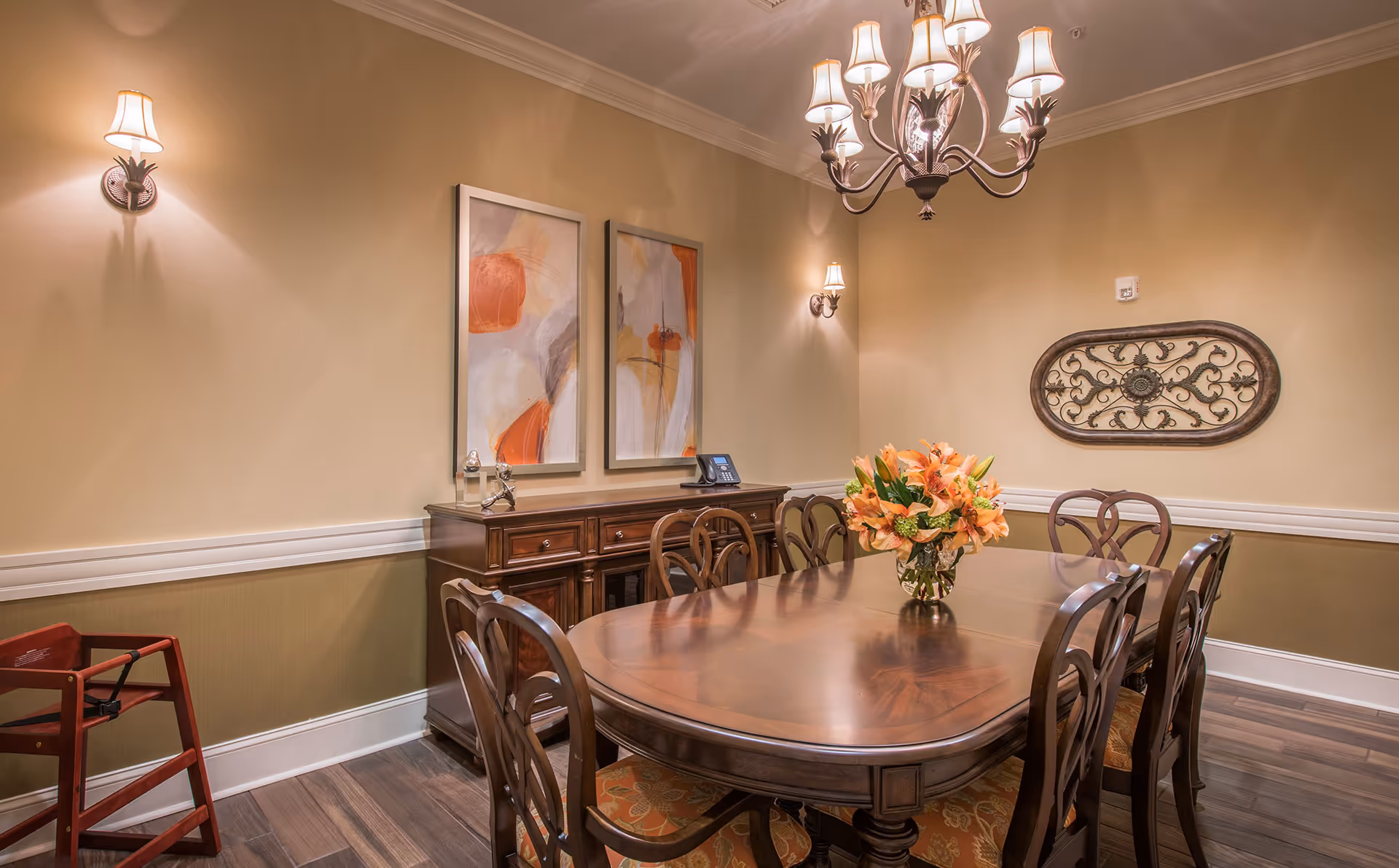 A warmly lit dining room with a polished wooden dining table surrounded by six matching chairs. A vase with orange flowers is centered on the table. The walls are painted beige with white trim and decorated with two abstract paintings and an ornate metal wall hanging. There is a wooden sideboard against the wall and a chandelier with multiple lampshades hanging above the table. A red wooden walker is positioned in the corner of the room.