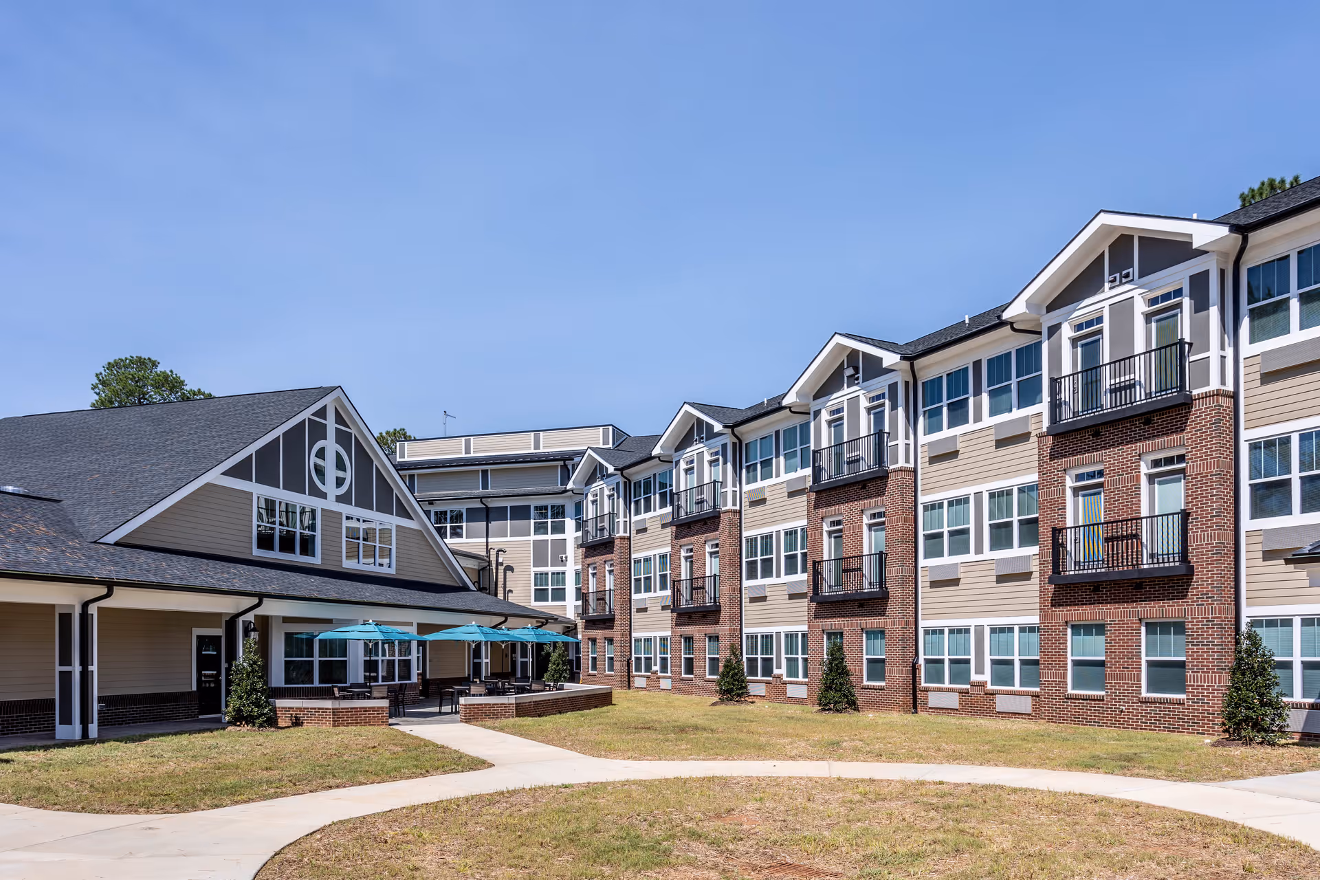 Exterior view of a senior living facility with a three-story building featuring multiple windows and small balconies. There is a covered patio area with tables and blue umbrellas, surrounded by a grassy courtyard and paved walkways under a clear blue sky.