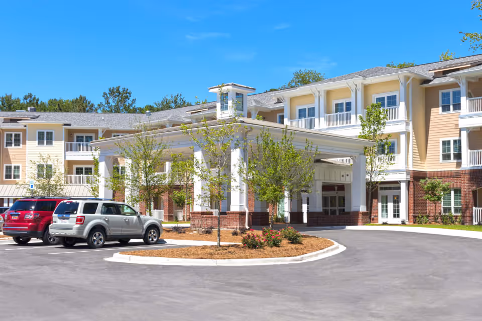 Exterior view of a senior living facility named Harmony at Wescott, showing a large covered entrance with white columns, a circular driveway, parked cars, and a multi-story building with balconies and windows under a clear blue sky.