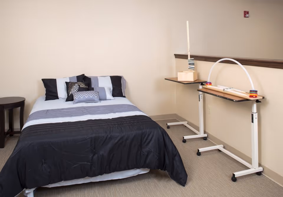 A small bedroom with a made bed, decorative pillows, a side table, and two overbed activity tables along a beige wall.