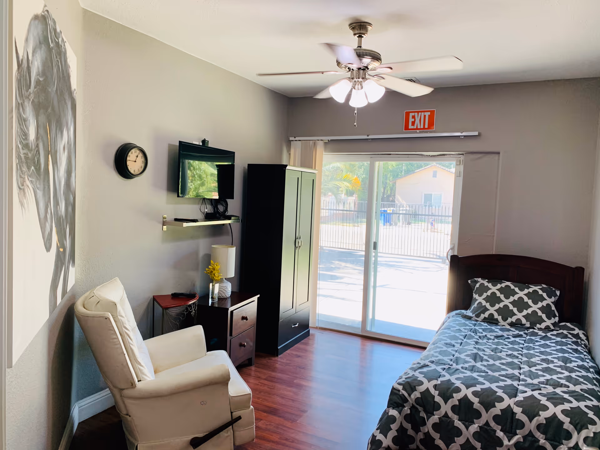 A small, neatly arranged bedroom with a single bed covered in a gray and white patterned comforter and matching pillow. There is a white recliner chair next to a small wooden nightstand with a lamp and a small vase with yellow flowers. A flat-screen TV is mounted on the wall above a small shelf. A large black wardrobe stands near sliding glass doors that lead outside. A ceiling fan with lights is mounted on the ceiling, and an exit sign is above the sliding doors. The walls are painted light gray, and there is a large black and white horse painting on one wall.