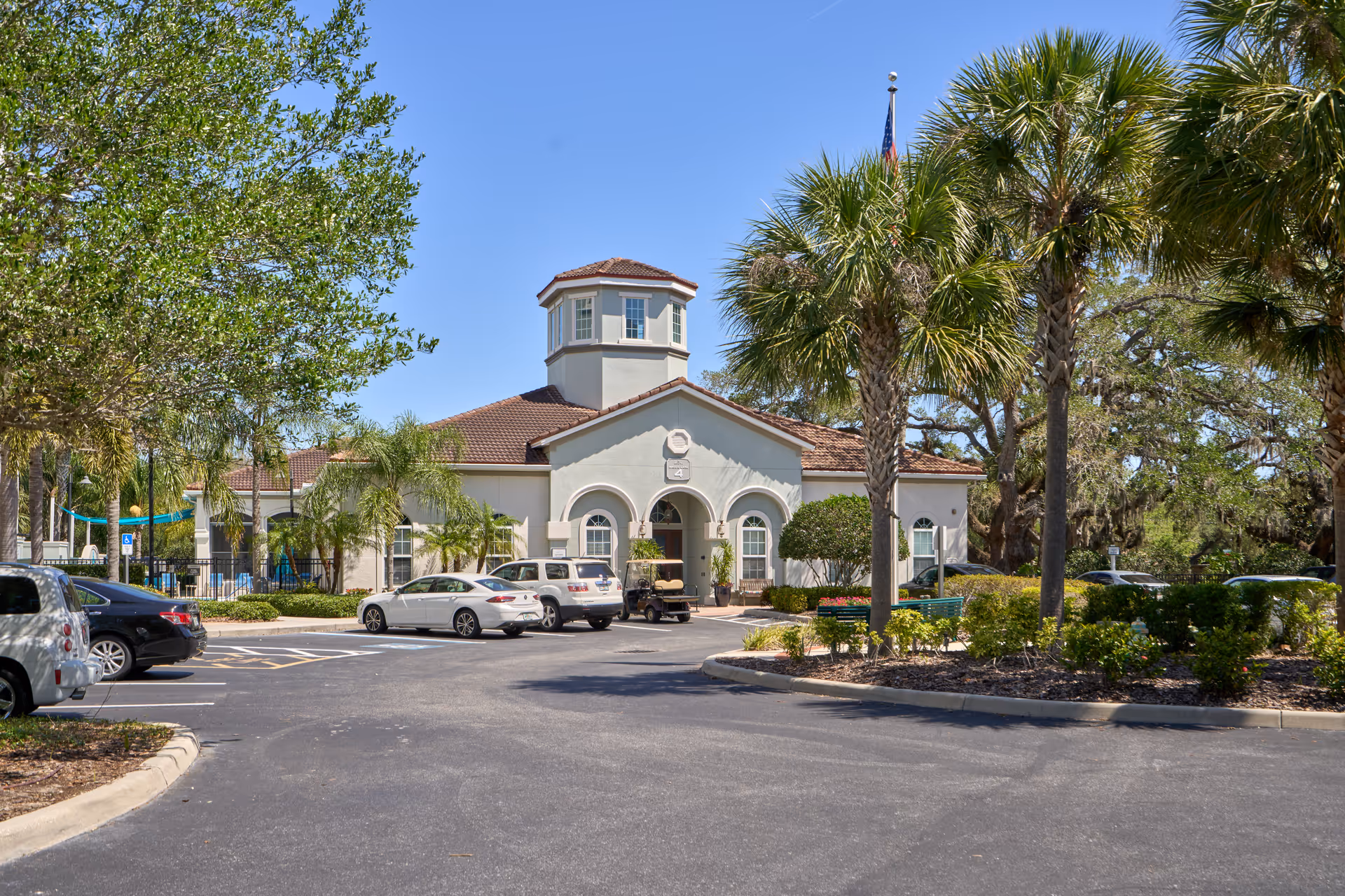 Exterior view of a single-story building with a tower-like structure on top, surrounded by palm trees and other greenery. Several cars are parked in front of the building under a clear blue sky.