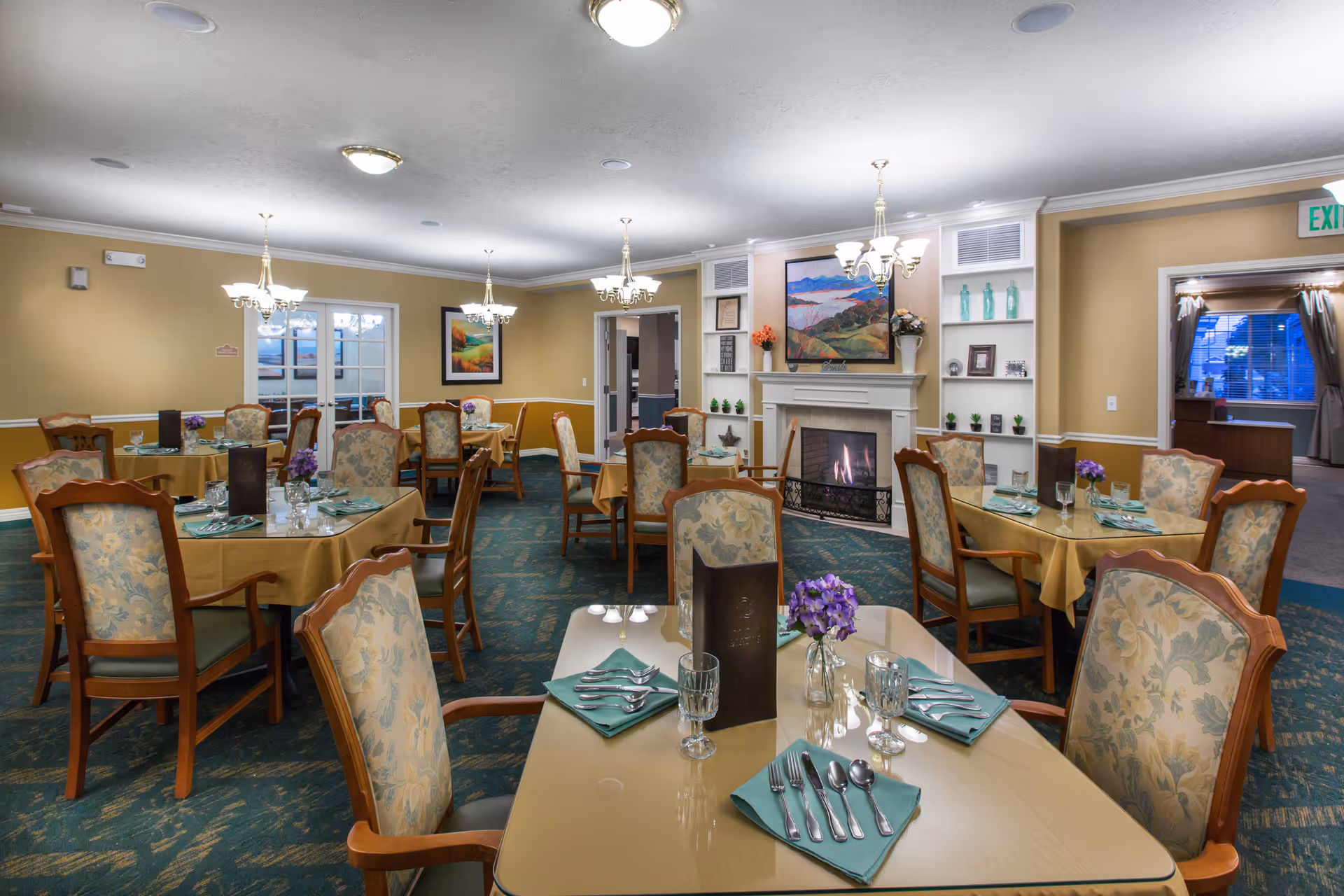 A cozy dining room in a senior living facility with several tables covered in yellow tablecloths, each set with green napkins, silverware, glasses, and small purple flower centerpieces. The room features upholstered wooden chairs, a fireplace with a painting above it, built-in shelves with decorative items, and warm lighting from chandeliers. There are doors and windows in the background, and an exit sign is visible.
