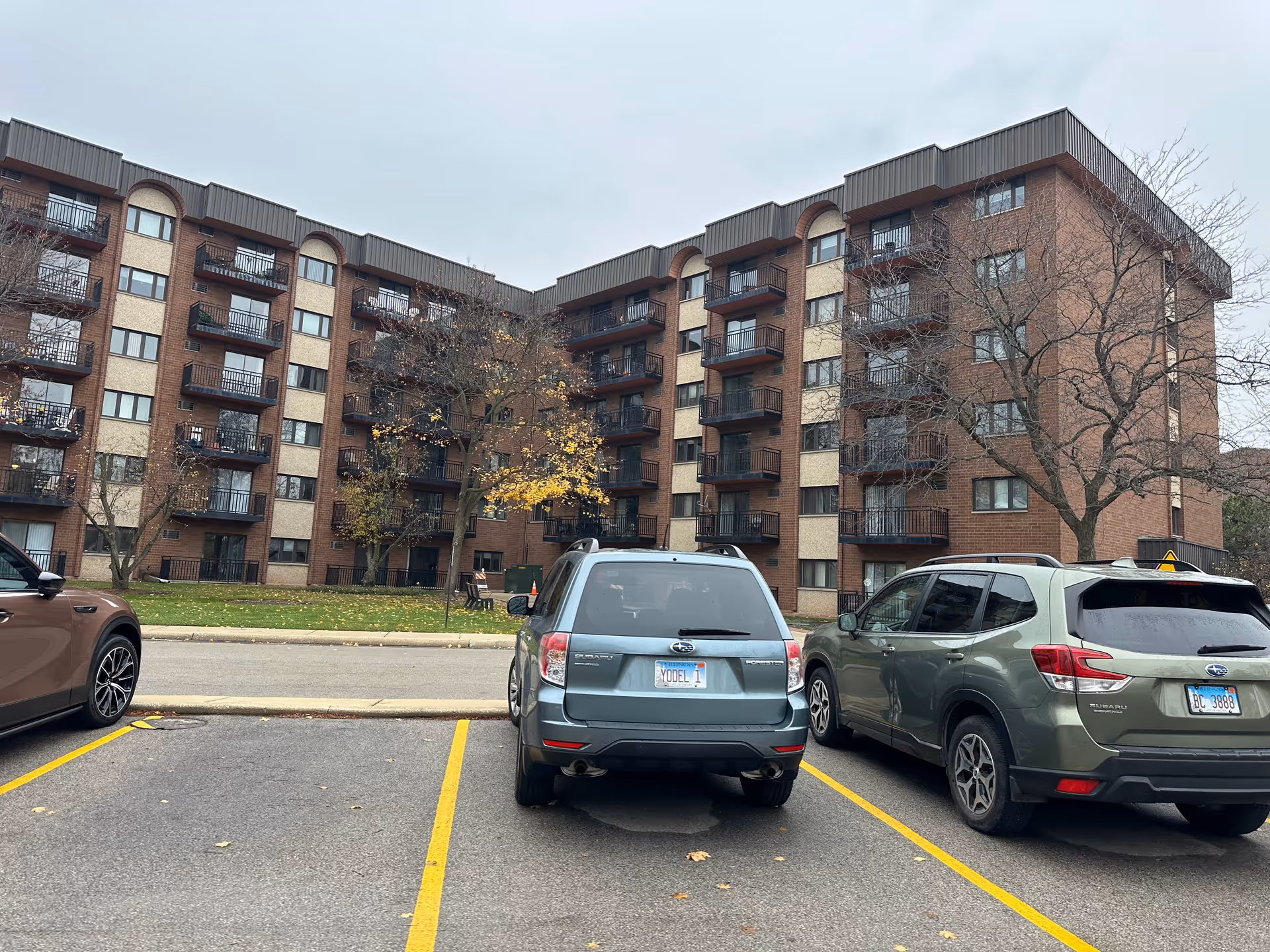 Exterior view of a multi-story brick residential building with balconies and several parked cars in the foreground under an overcast sky.