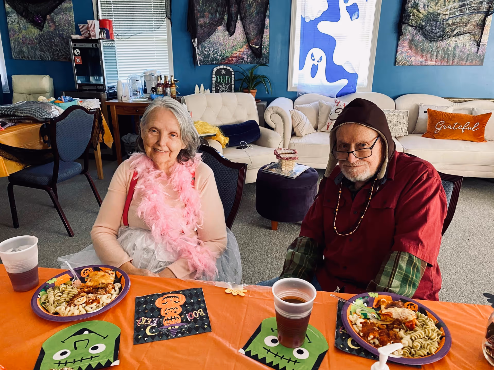 Two elderly people sitting at a table with Halloween-themed decorations and plates of food in a cozy living room. The woman on the left is wearing a pink feather boa and a light-colored outfit, while the man on the right is wearing glasses, a red shirt, and a hood. Behind them are sofas with decorative pillows, including one that says 'Grateful', and Halloween decorations on the windows and walls.