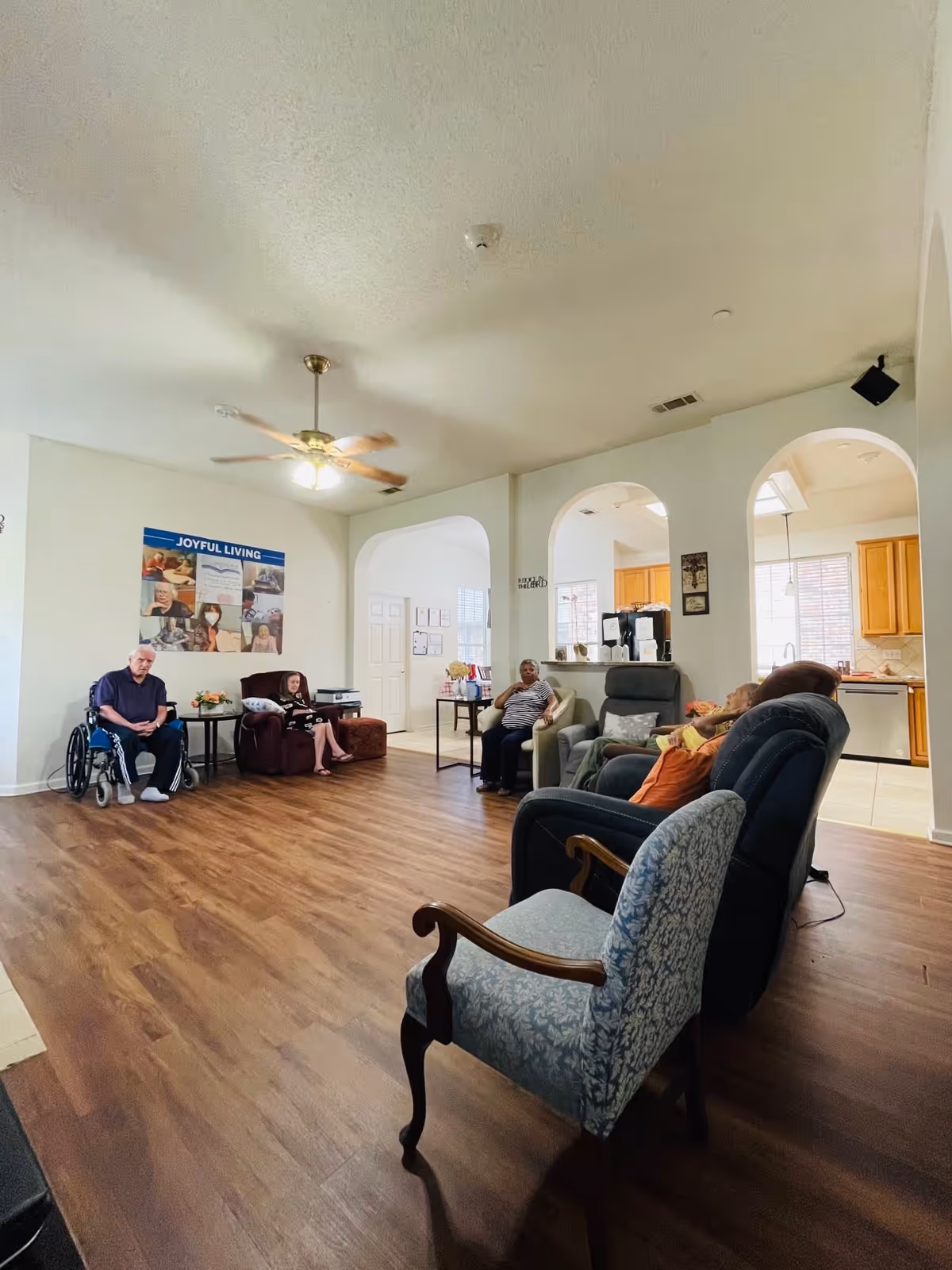 A spacious living room in an assisted living facility with wooden flooring and a ceiling fan. Several elderly residents are seated on various chairs and recliners arranged around the room. The room has white walls with a poster titled 'Joyful Living' and an open view into a kitchen area with wooden cabinets and windows letting in natural light.