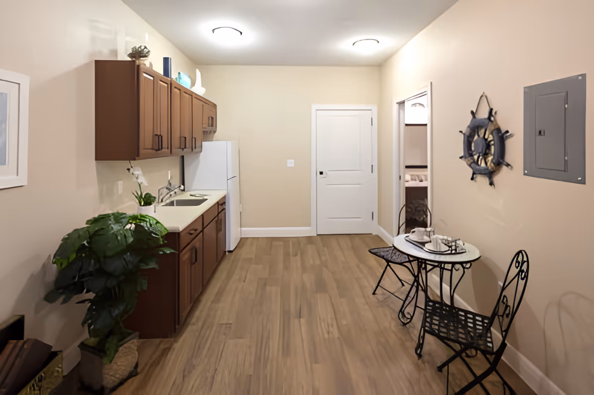 A small kitchen area with wooden cabinets, a white refrigerator, and a sink on the left side. On the right side, there is a small round table with two black metal chairs. A decorative mirror shaped like a ship's wheel hangs on the wall above the table. The floor is wooden, and the walls are painted beige. There is a potted plant on the floor near the cabinets and a white door at the end of the room.