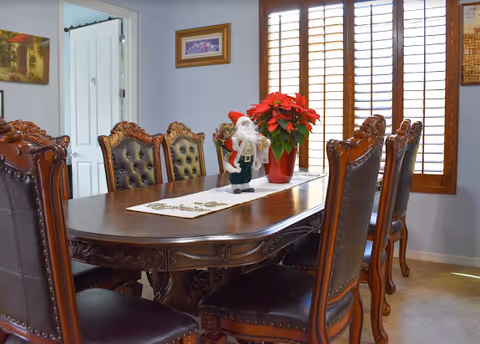 A dining room with a large wooden dining table surrounded by eight ornate wooden chairs with leather upholstery. On the table, there is a white table runner with a small Santa Claus figurine and a red poinsettia plant. The room has light-colored walls, a window with wooden shutters, and framed artwork on the walls.