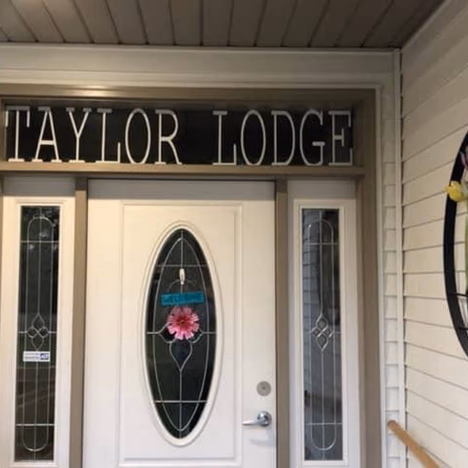 Front entrance door of Taylor Lodge with decorative glass panels on the door and sidelights, a pink flower decoration hanging on the door, and the name 'TAYLOR LODGE' displayed above the door.