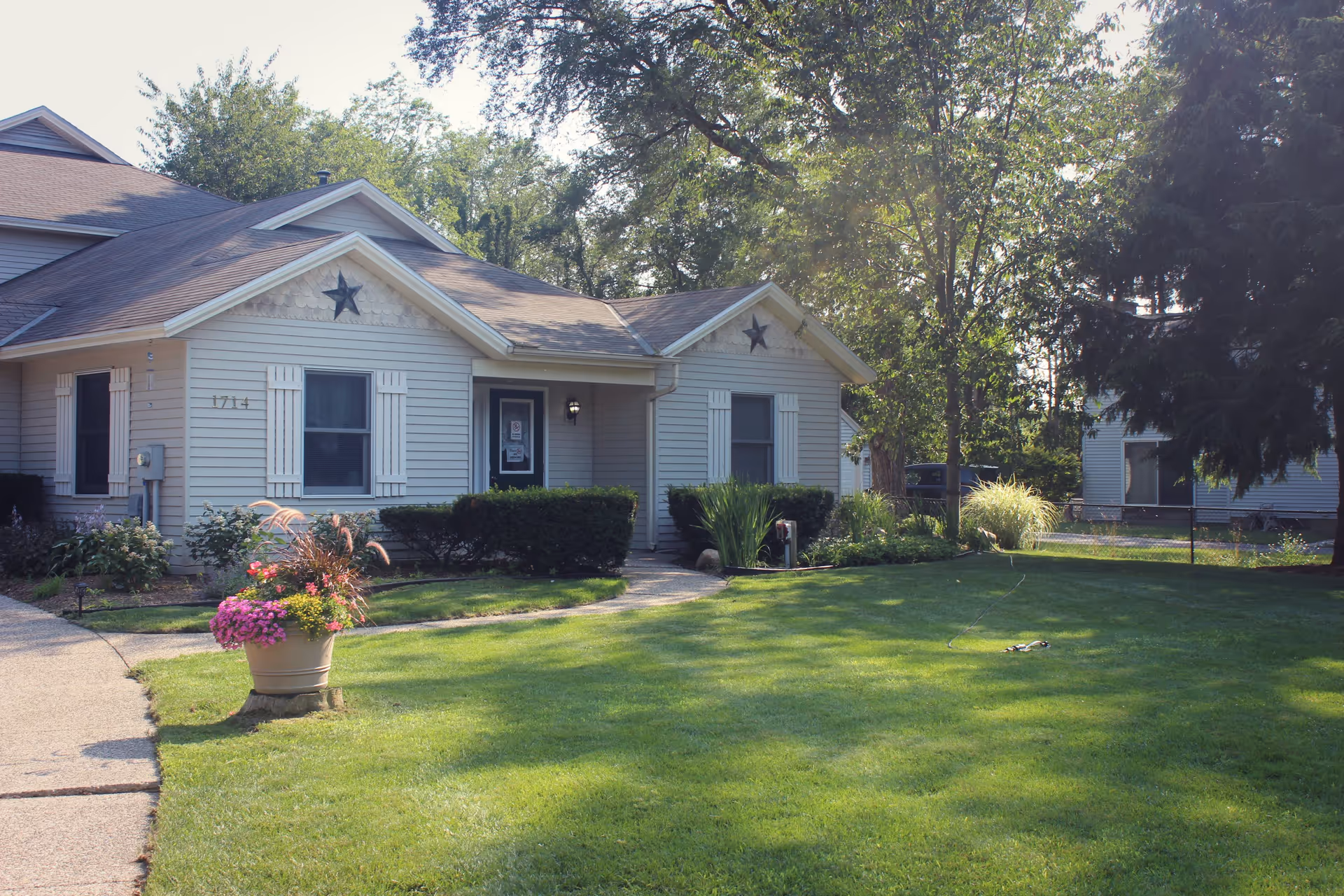 Exterior view of a single-story residential building with beige siding, two windows with white shutters, and a front door under a small porch. The building has two decorative stars on the gables. There is a well-maintained green lawn with a flower pot containing pink and green plants near a curved concrete walkway. Trees and bushes surround the building under a sunny sky.