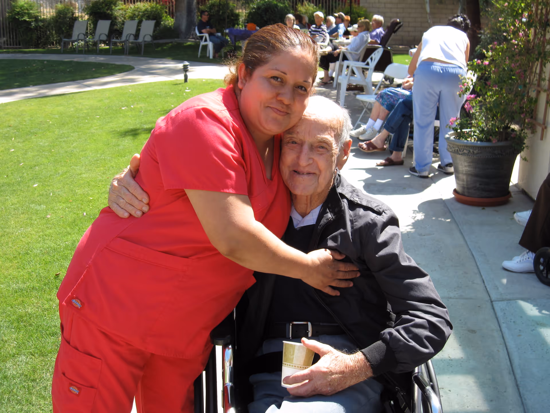 A caregiver in red scrubs hugging an elderly man sitting in a wheelchair outdoors on a sunny day. In the background, several elderly people are seated on white chairs along a walkway next to a building, with green grass and trees nearby.