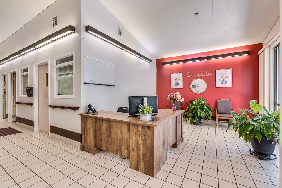 Reception area with a wooden desk, tiled floor, potted plants, seating, and a red accent wall bearing the phrase 'Love is spoken here'.