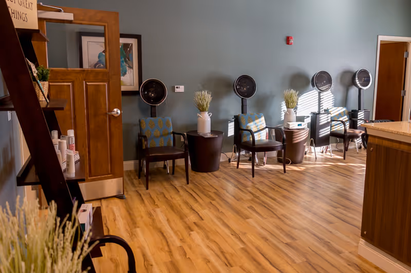 Interior of a salon area with four vintage hair dryers positioned above chairs with patterned cushions. The room has wooden flooring, a teal-colored wall, and wooden doors. There are small tables with white vases holding decorative plants between the chairs. A wooden shelf with hair care products is visible on the left side.