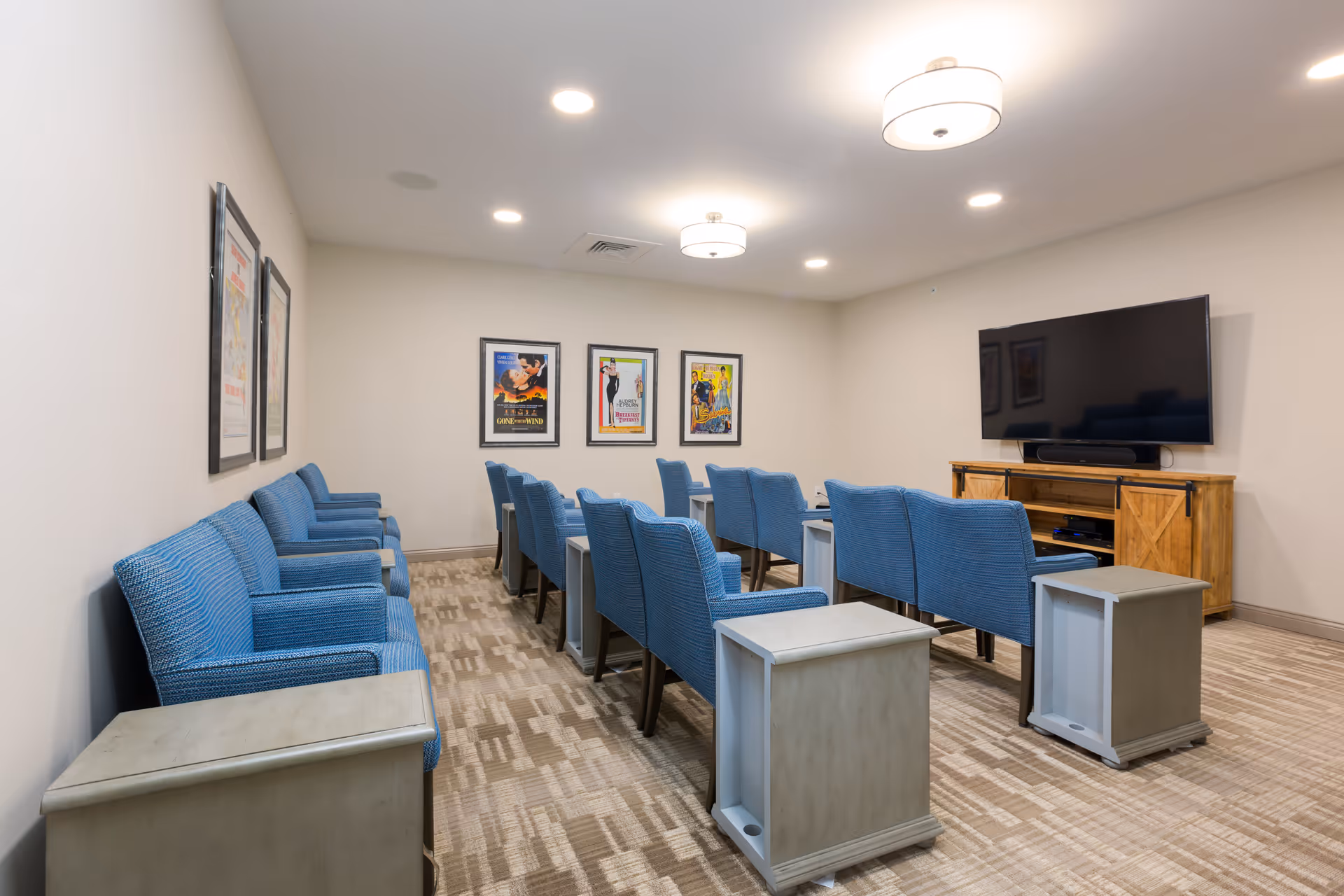A small theater-style room with rows of blue upholstered chairs facing a large flat-screen TV mounted on a wooden cabinet. The room has beige walls, carpeted floor, ceiling lights, and framed movie posters on the walls.