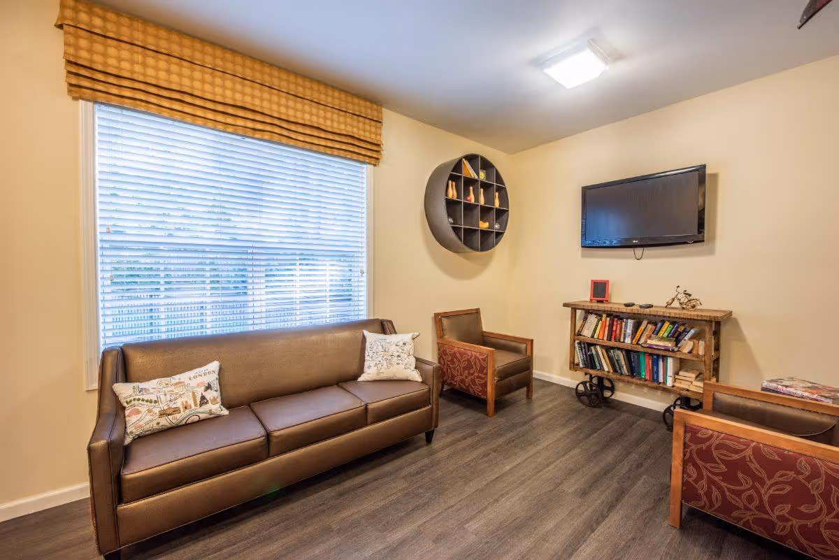 A cozy living room with a brown leather sofa adorned with two decorative pillows, two armchairs with patterned upholstery, a wall-mounted flat-screen TV, a rustic bookshelf filled with books, and a round wall shelf with decorative items. The room has a large window with blinds and a yellowish-beige wall color.