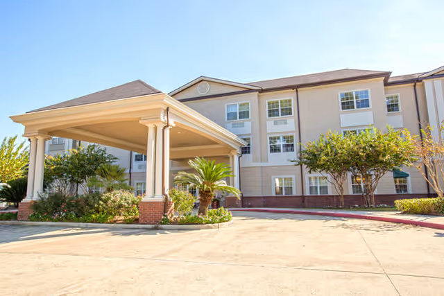Exterior view of a multi-story senior living facility building with a covered entrance supported by columns, surrounded by landscaped greenery and trees under a clear blue sky.
