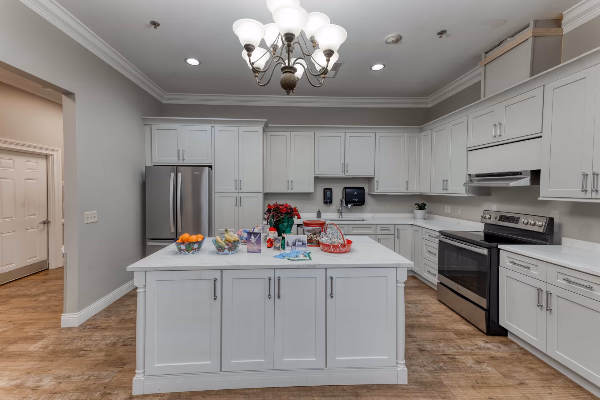 A spacious kitchen with white cabinets and a large white island in the center. The island has bowls of fruit, holiday cards, a poinsettia plant, and festive decorations on top. The kitchen features stainless steel appliances including a refrigerator and an oven with a range hood. The floor is wood, and the ceiling has recessed lighting and a chandelier.