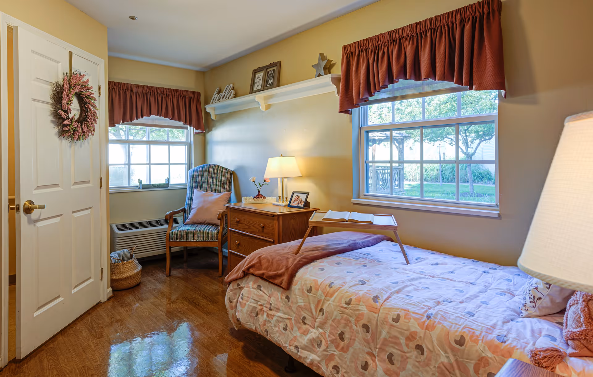 A cozy bedroom with a single bed covered in a patterned bedspread and a brown throw blanket. There is a wooden tray with an open book on the bed. Next to the bed is a wooden nightstand with a lamp, a small flower vase, and a framed photo. A cushioned armchair with a pink pillow is placed near two windows with red valances, letting in natural light and showing a view of greenery outside. A door with a decorative wreath is visible on the left side of the room, and the floor is polished wood.